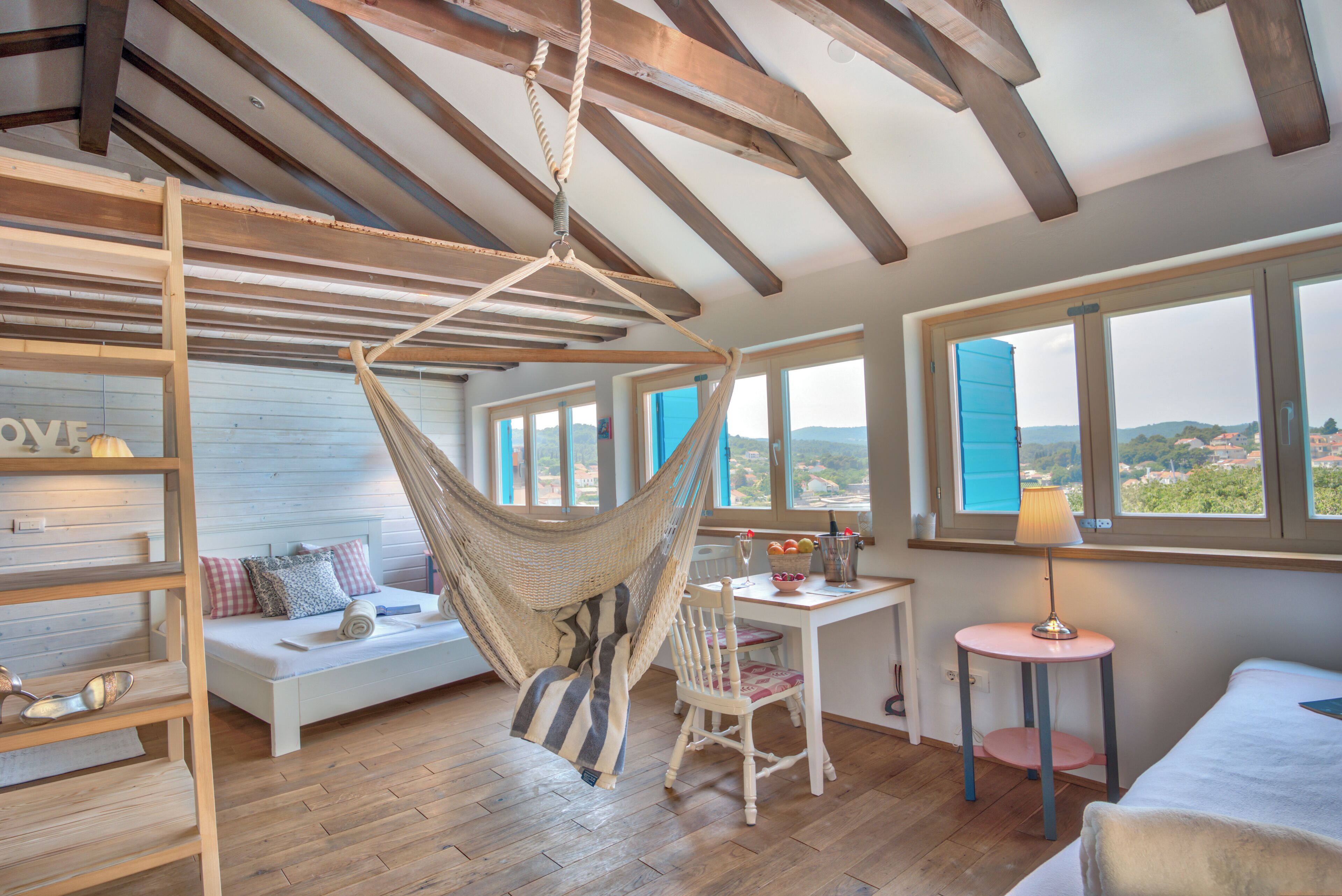A cozy room with a hammock, a wooden ceiling, a bed, a desk with a lamp, and large windows offering a view.