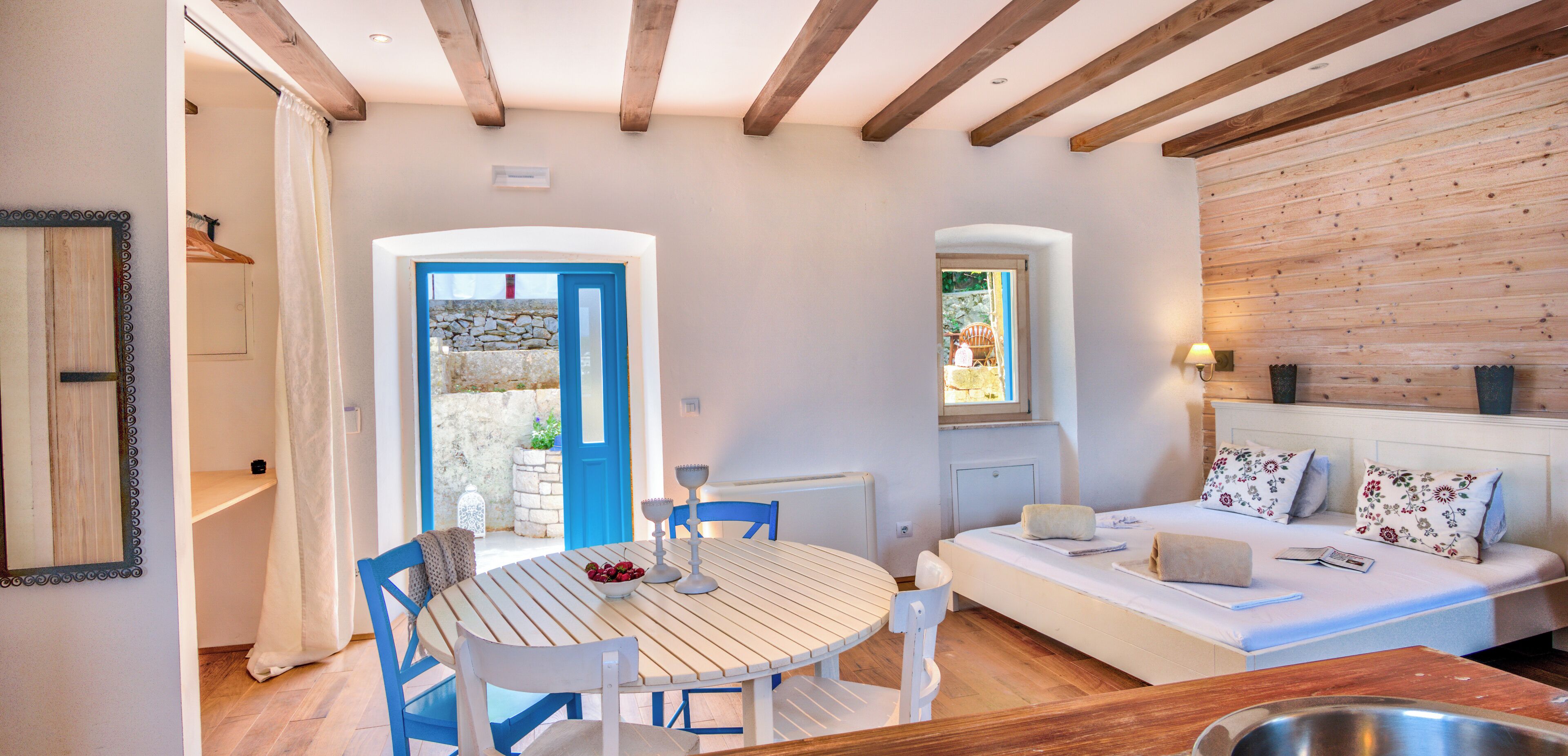 A cozy room with a wooden ceiling, a white sofa with floral pillows, a round wooden table with chairs, and a stone wall visible through an open door.