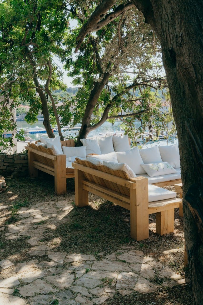 Two wooden benches with white cushions, positioned under a tree, overlooking a body of water.