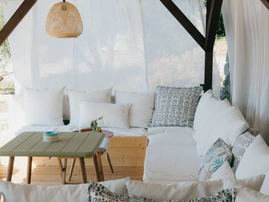 A covered outdoor seating area with a wooden table, white cushions, and a woven hanging light fixture.