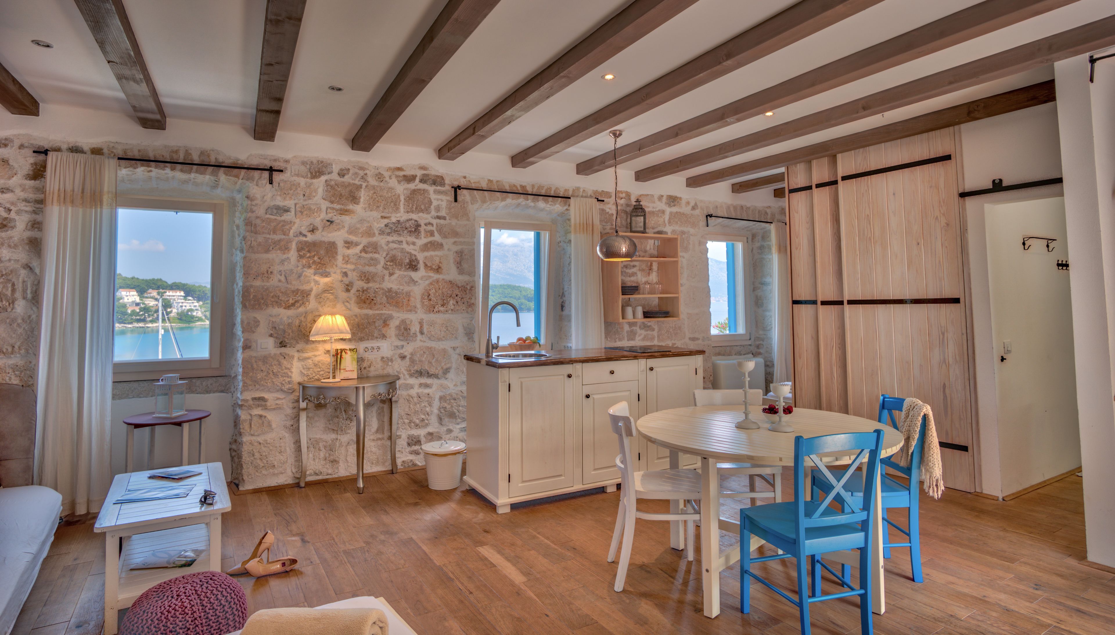 A cozy kitchen with a stone wall, wooden beams, a dining area with blue chairs, and a view of the sea through large windows.