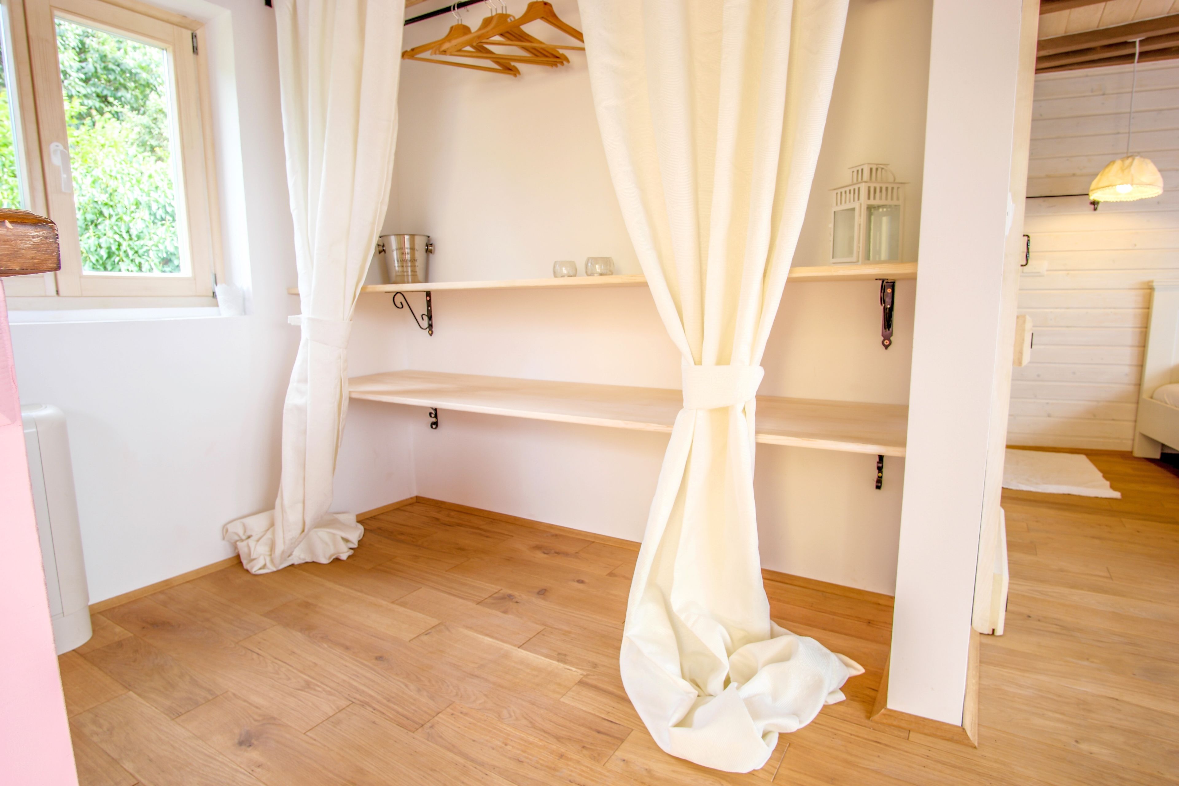 Une pièce avec un plancher en bois, une fenêtre donnant sur un paysage verdoyant, un rideau blanc et une étagère sur laquelle est suspendu un luminaire.