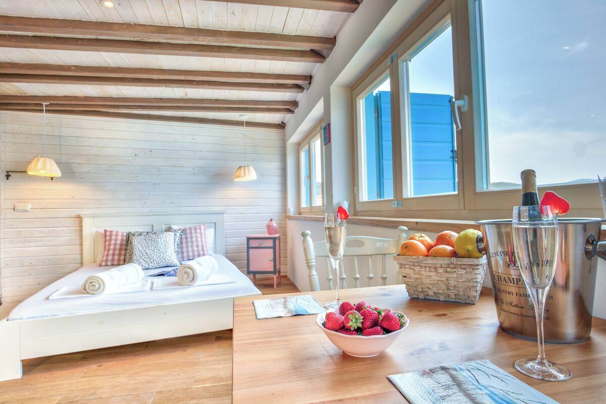 Une chambre confortable avec un plafond en bois, un lit avec des oreillers, une table de chevet sur laquelle se trouve un bol de fraises, un seau à champagne et une fenêtre donnant sur un beau paysage.
