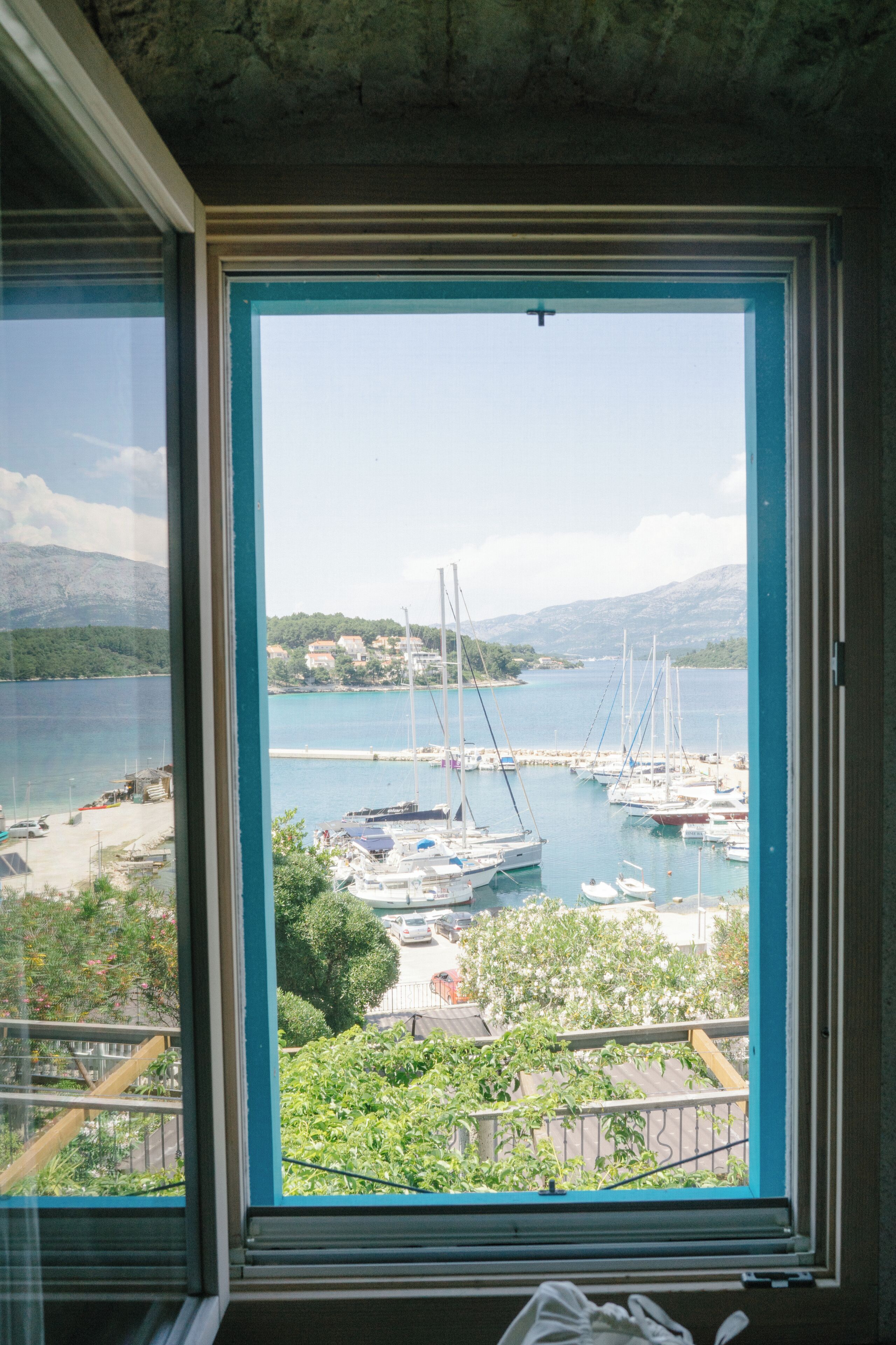 Une vue depuis une fenêtre donnant sur une marina, avec des bateaux, une plage et de la verdure.