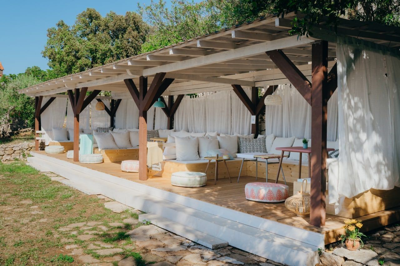 An outdoor covered patio with seating and tables, surrounded by a stone wall and greenery.