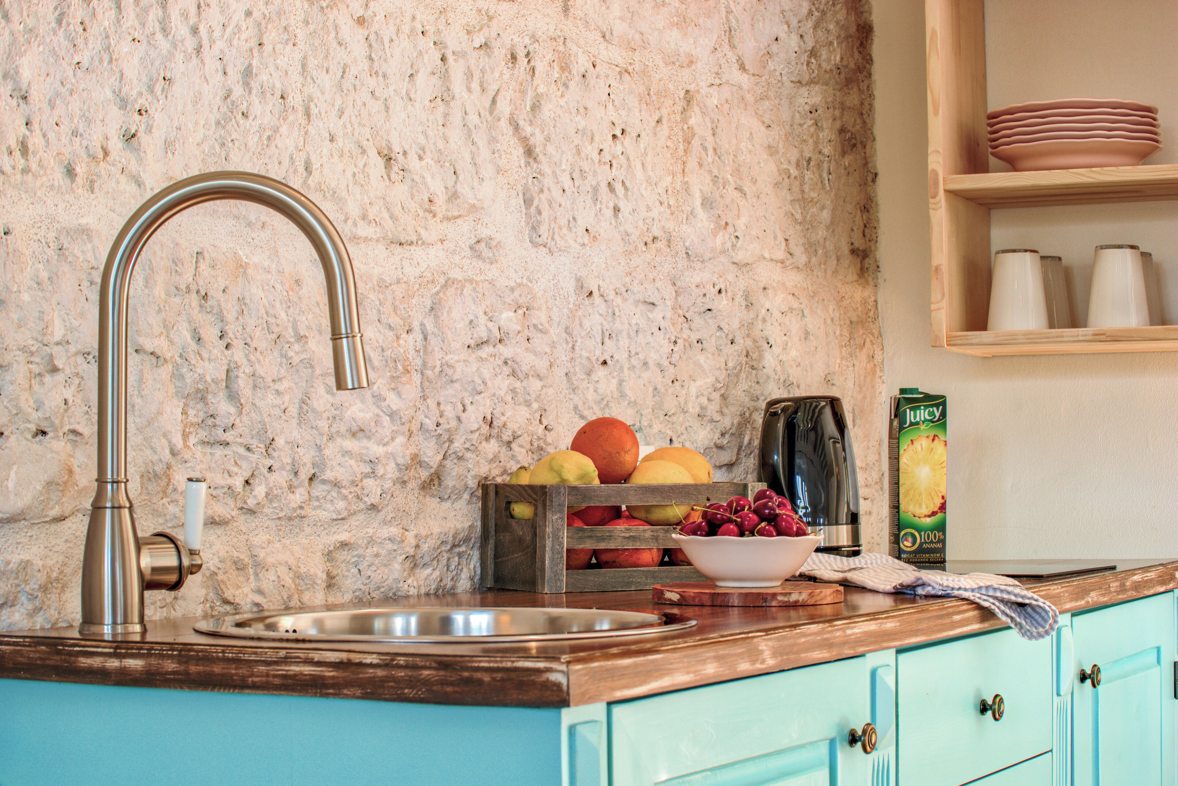 A kitchen with a stone wall, a stainless steel sink, a wooden countertop, and a fruit crate.