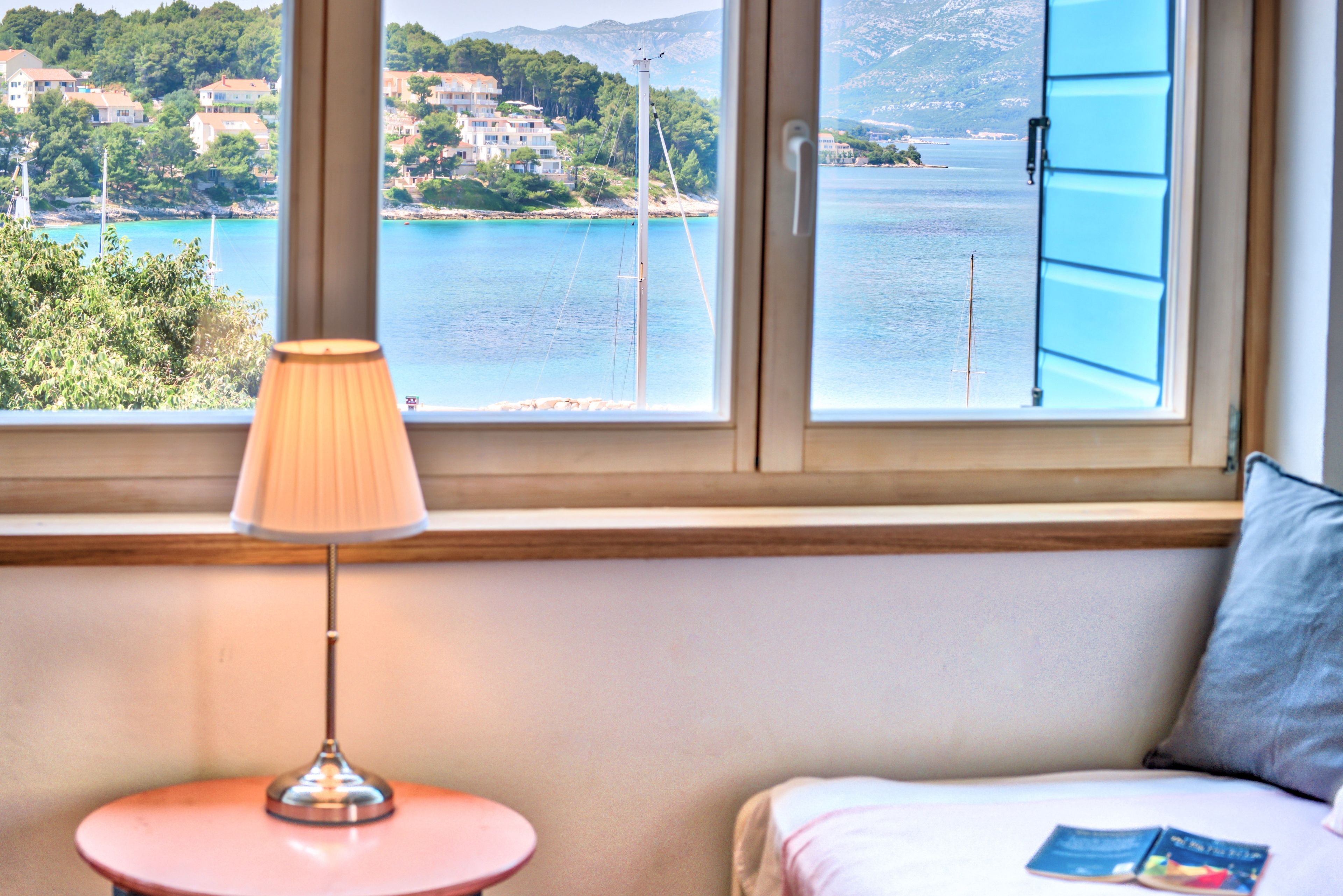 Una habitación con vista a la costa, una lámpara sobre una mesita y una cama con un cojín y un libro.