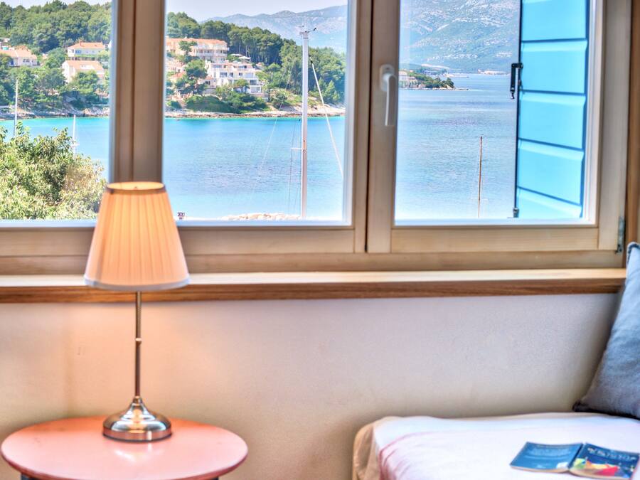 Una habitación con vista a la costa, una lámpara sobre una mesita y una cama con un cojín y un libro.