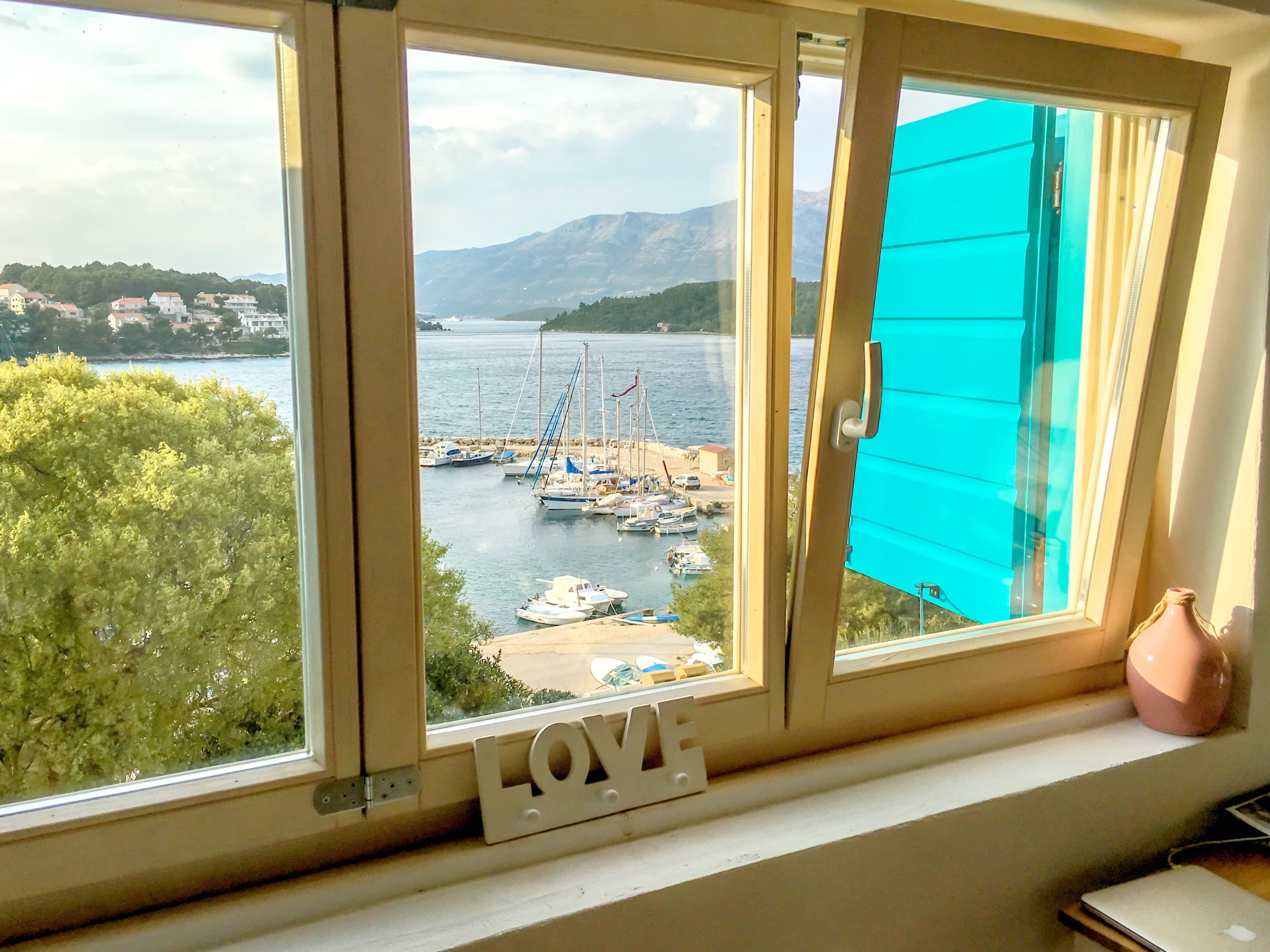 Una ventana con vista a un puerto deportivo, barcos y un paisaje montañoso.