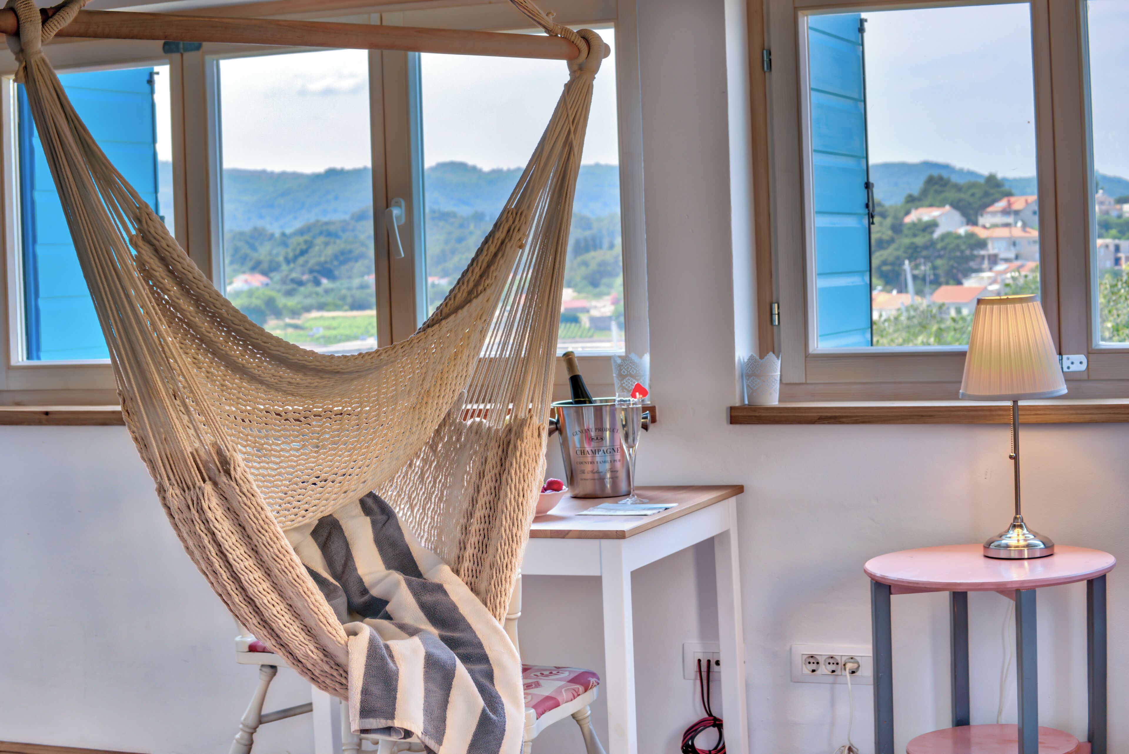 Una habitación con una silla hamaca, un escritorio con lámpara y vista a un pueblo a través de ventanas abiertas.