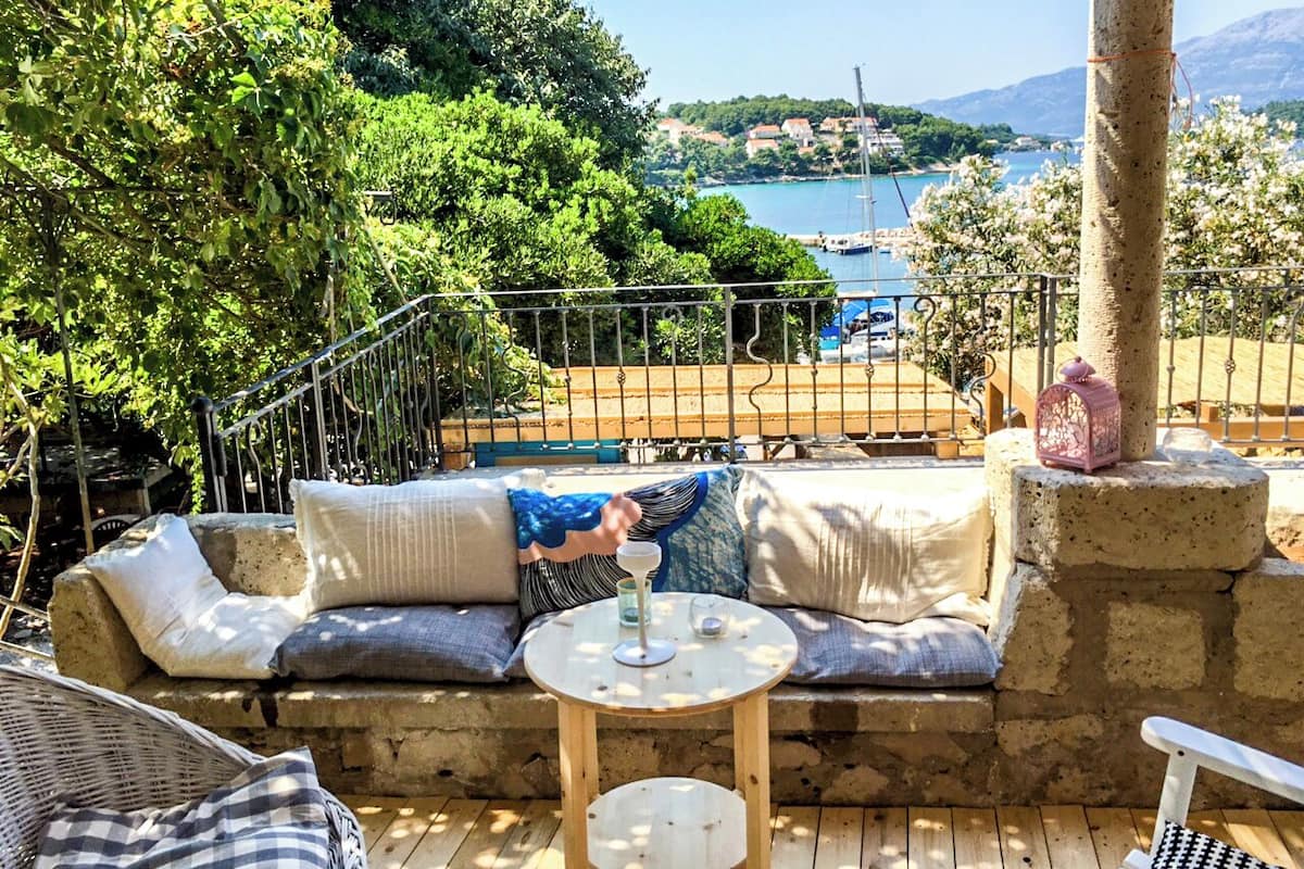 Une terrasse avec un banc en pierre, une petite table ronde et un coin salon agrémenté de coussins, offrant une vue sur une étendue d’eau et un paysage montagneux.