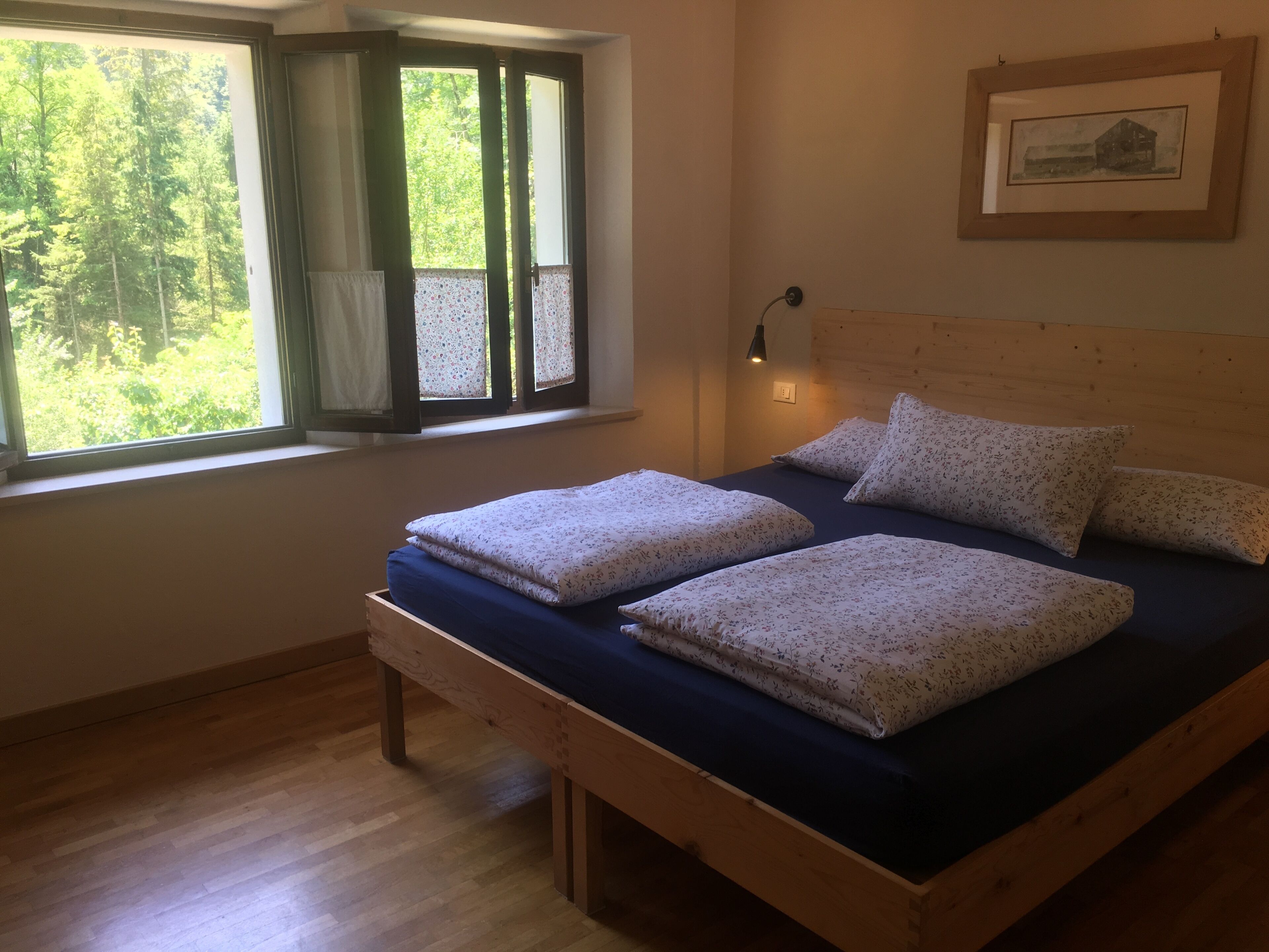 Un dormitorio con cama de madera, una ventana con cortinas, un cuadro enmarcado en la pared y una lámpara de noche.