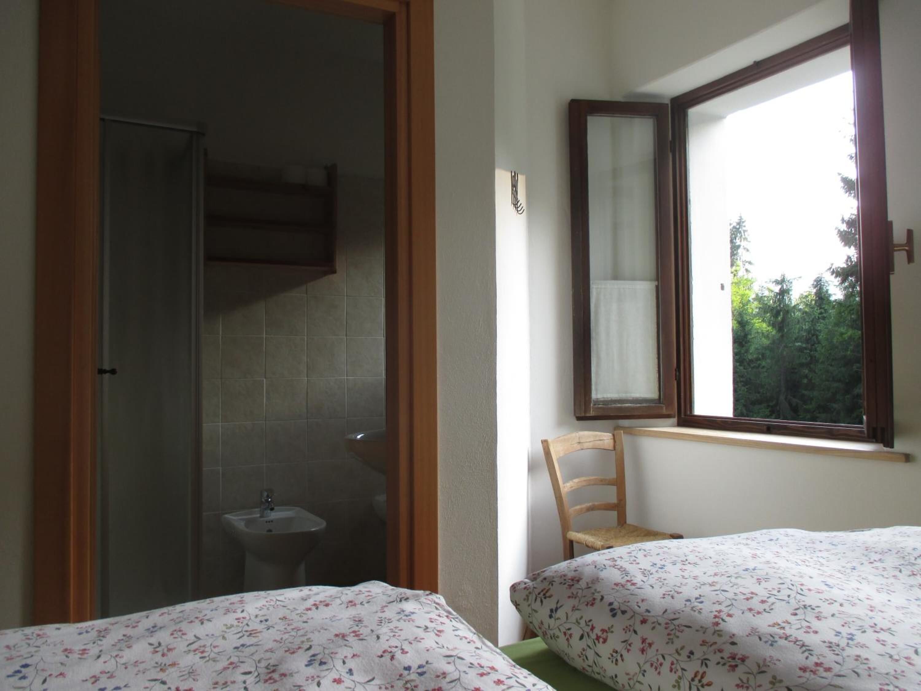 A bedroom with two beds, a window with a view of trees, a wooden chair, and a bathroom with a shower and sink.