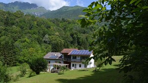 A two-story house with a sloped roof and solar panels, surrounded by lush greenery and mountains in the background. - Alla Casetta (Cesiomaggiore)