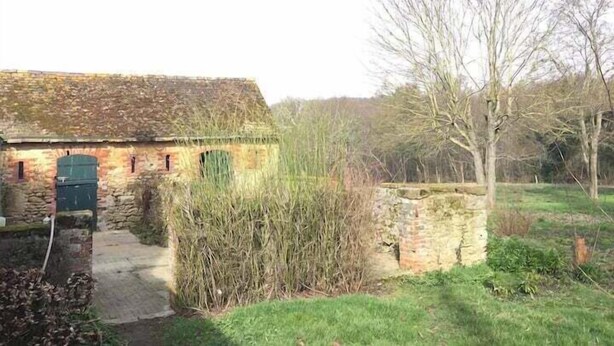 Een landelijk stenen gebouw met schuine kap, een smalle deuropening en een kleine tuin met gras en struiken.