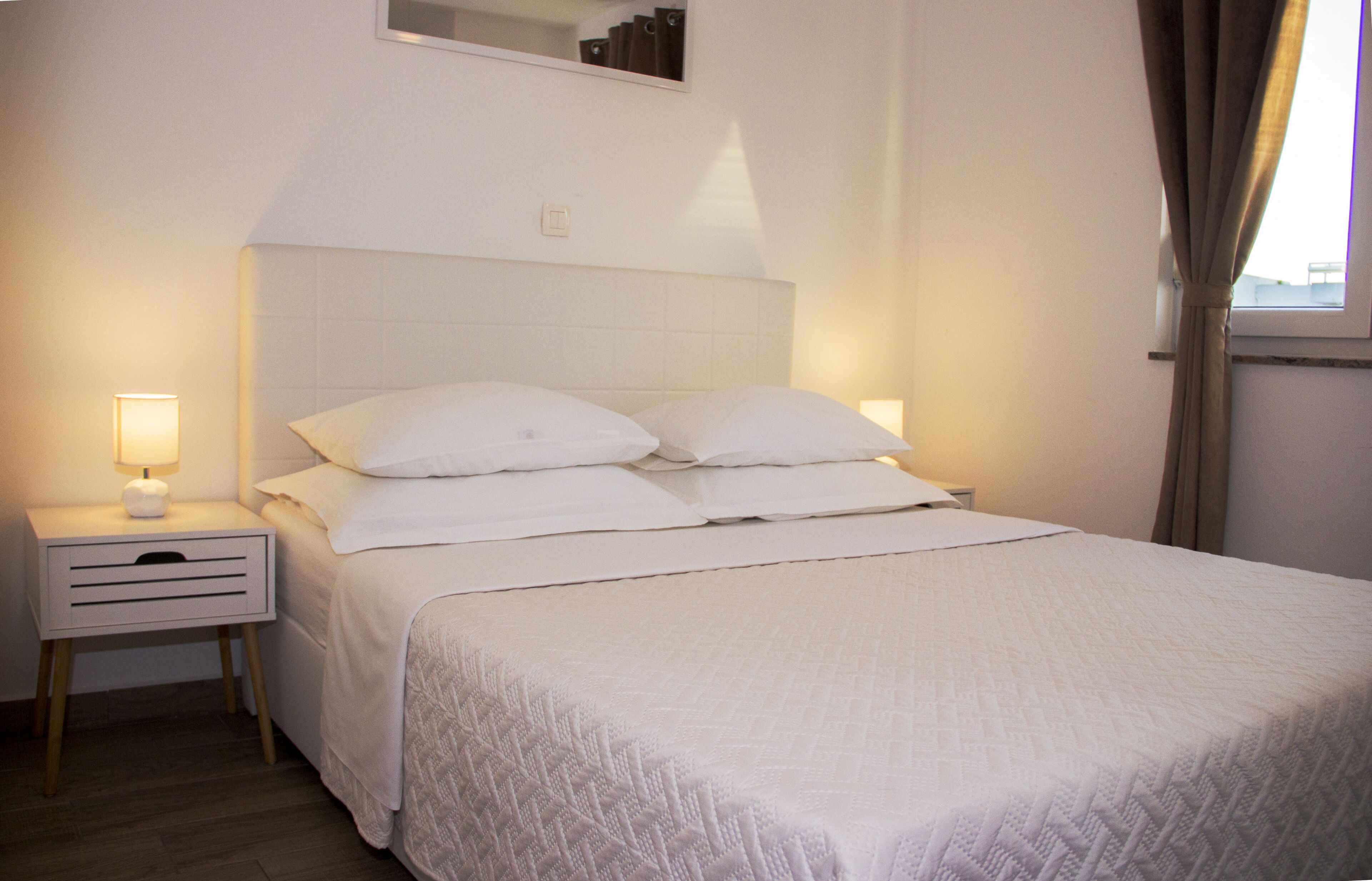 Una cama bien tendida con sábanas blancas y un cabecero, una mesita de noche con una lámpara, y una ventana con cortinas.