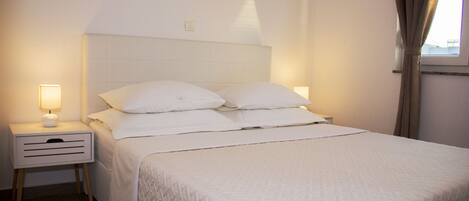 Una cama bien tendida con sábanas blancas y un cabecero, una mesita de noche con una lámpara, y una ventana con cortinas.