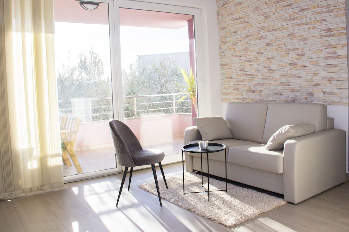 A modern living room with a sofa, a chair, a small table, and a view through a sliding glass door to an outdoor balcony.