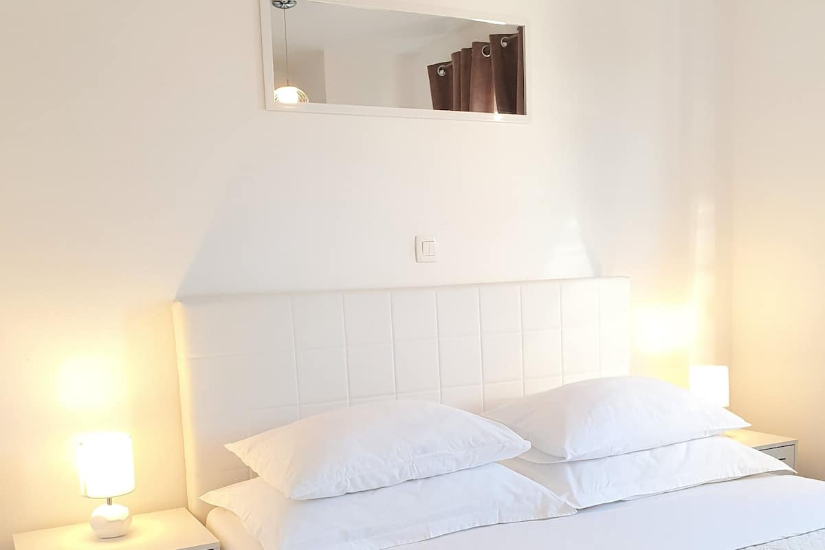 A neatly made bed with white bedding, a bedside table with a lamp, and a wall-mounted mirror.