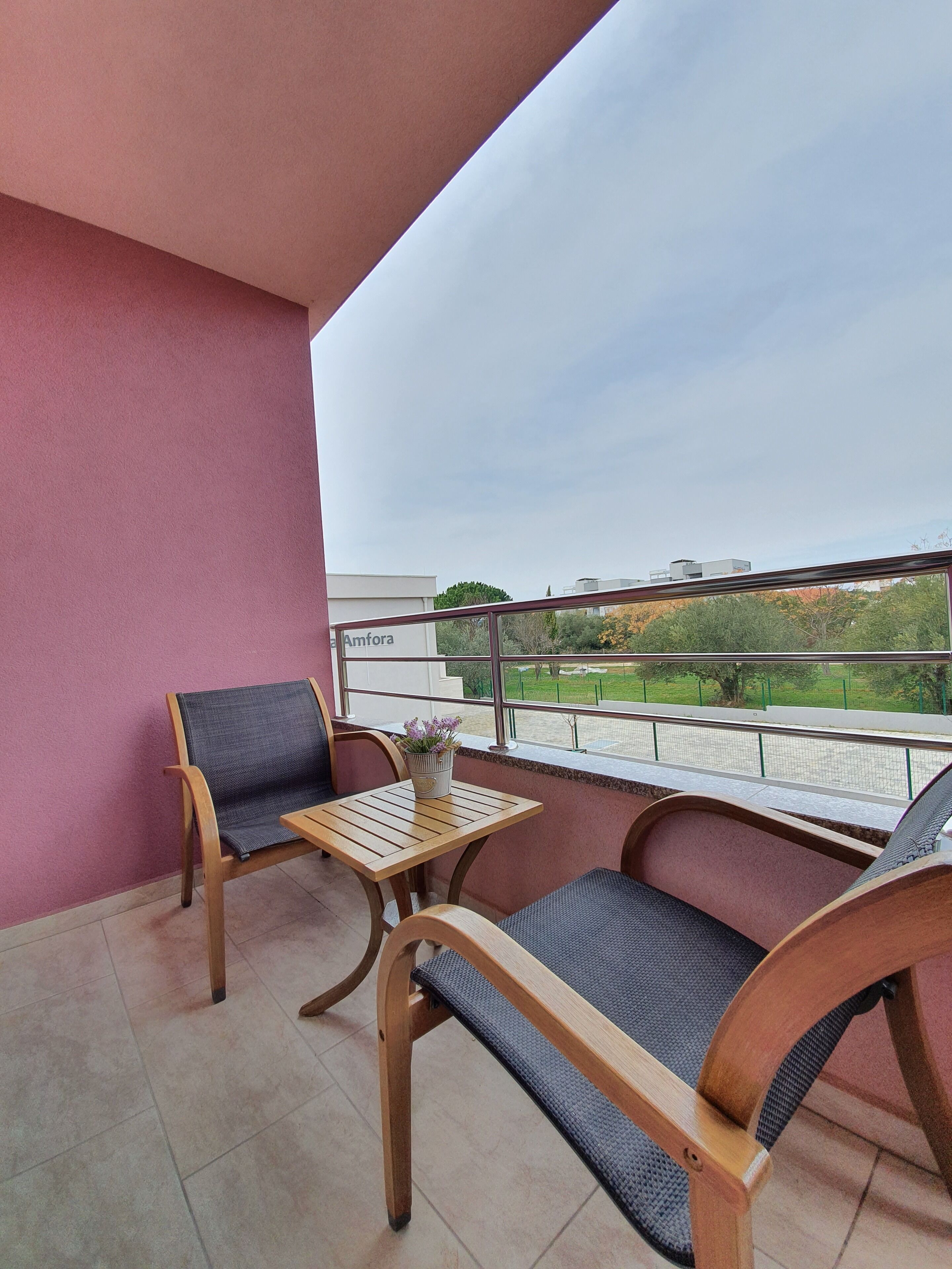 Ein Balkon mit zwei Holzstühlen, einem kleinen Tisch, einem Blumentopf und Blick auf einen grünen Park sowie Gebäude.