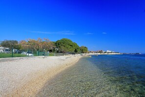 A pebble beach with clear water, a large tree, and buildings in the background. - Apartments Karla (Bibinje)