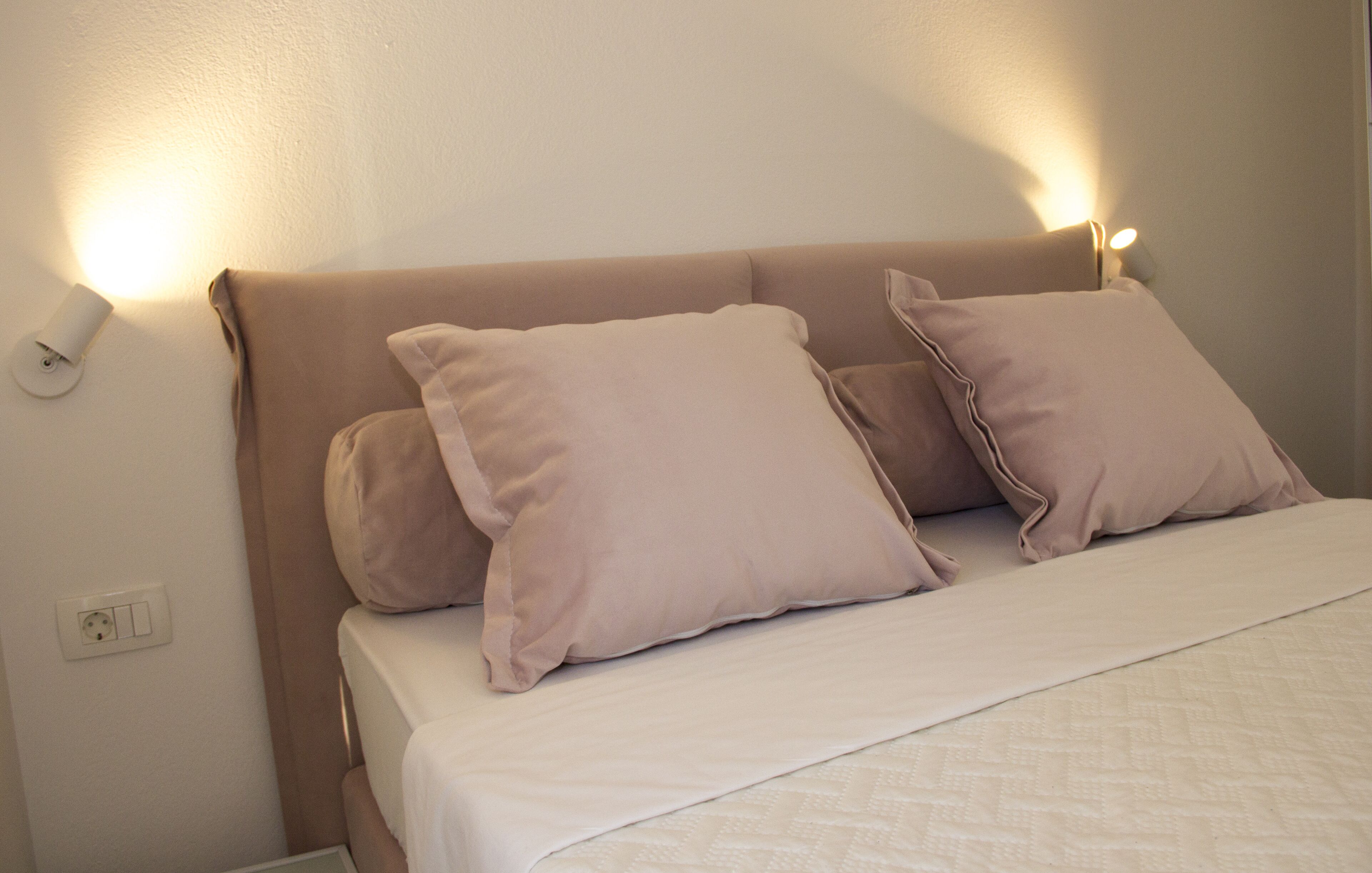 A bed with two pillows and a white blanket, a wall socket, and a wall-mounted light fixture.