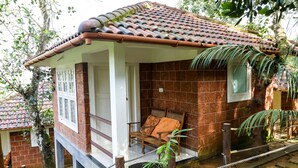 A small brick house with a tiled roof, a porch with a bench, and a wooden fence. - Wayanad Ranches Resorts (Vythiri)