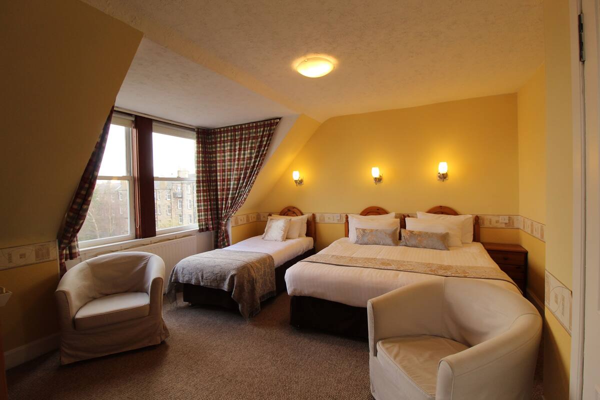 A hotel room with two single beds, two armchairs, a window with curtains, and wall-mounted lights.