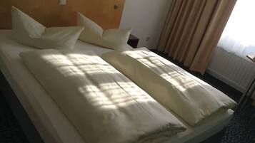 A neatly made bed with white linens and pillows, a wooden headboard, and a window with curtains.