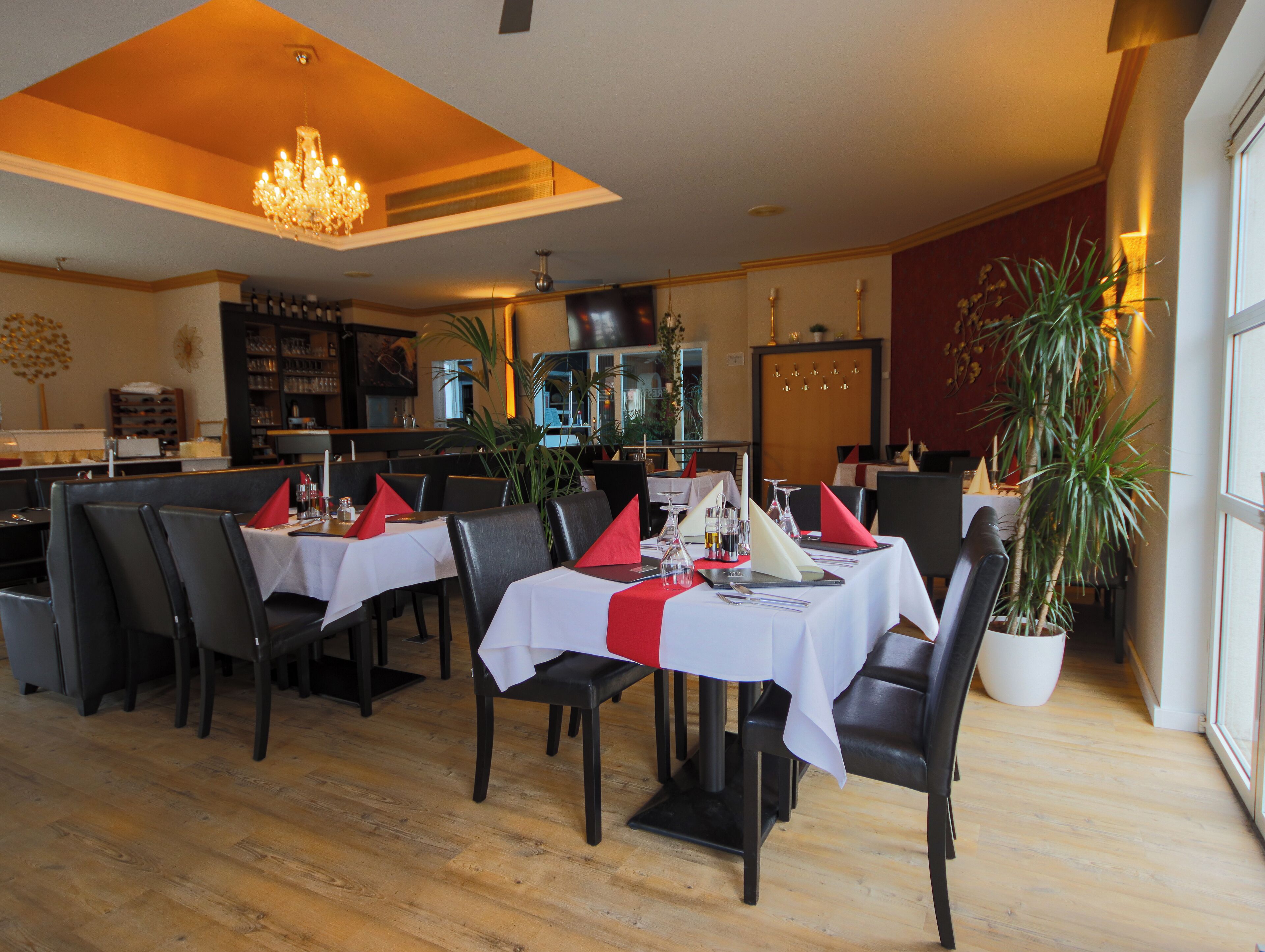 A restaurant with tables set for dining, chairs, a chandelier, and a potted plant.