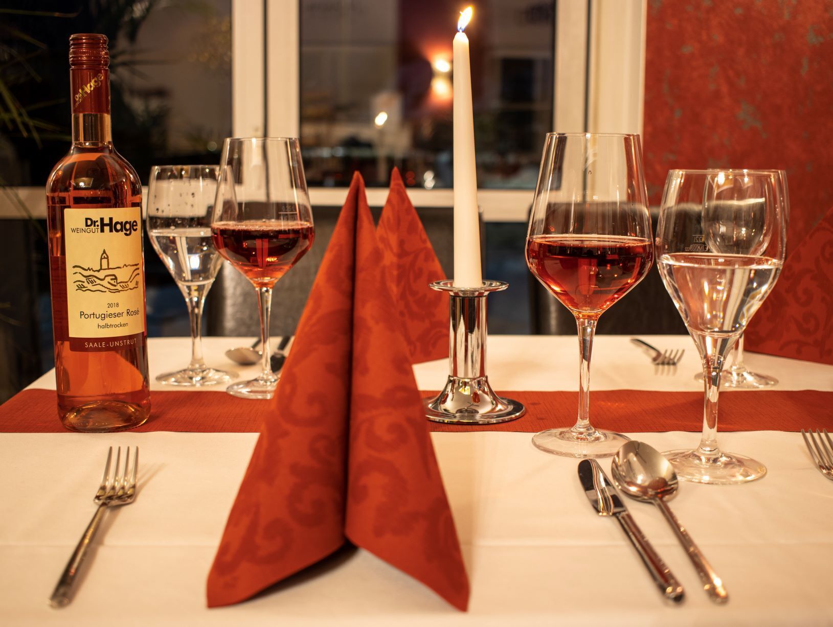 A table set with wine glasses, a bottle of Dr. Hage wine, and a lit candle.