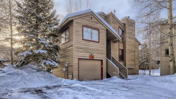 A two-story wooden house with a garage, surrounded by snow-covered trees.