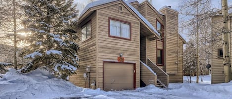 A two-story wooden house with a garage, surrounded by snow-covered trees.