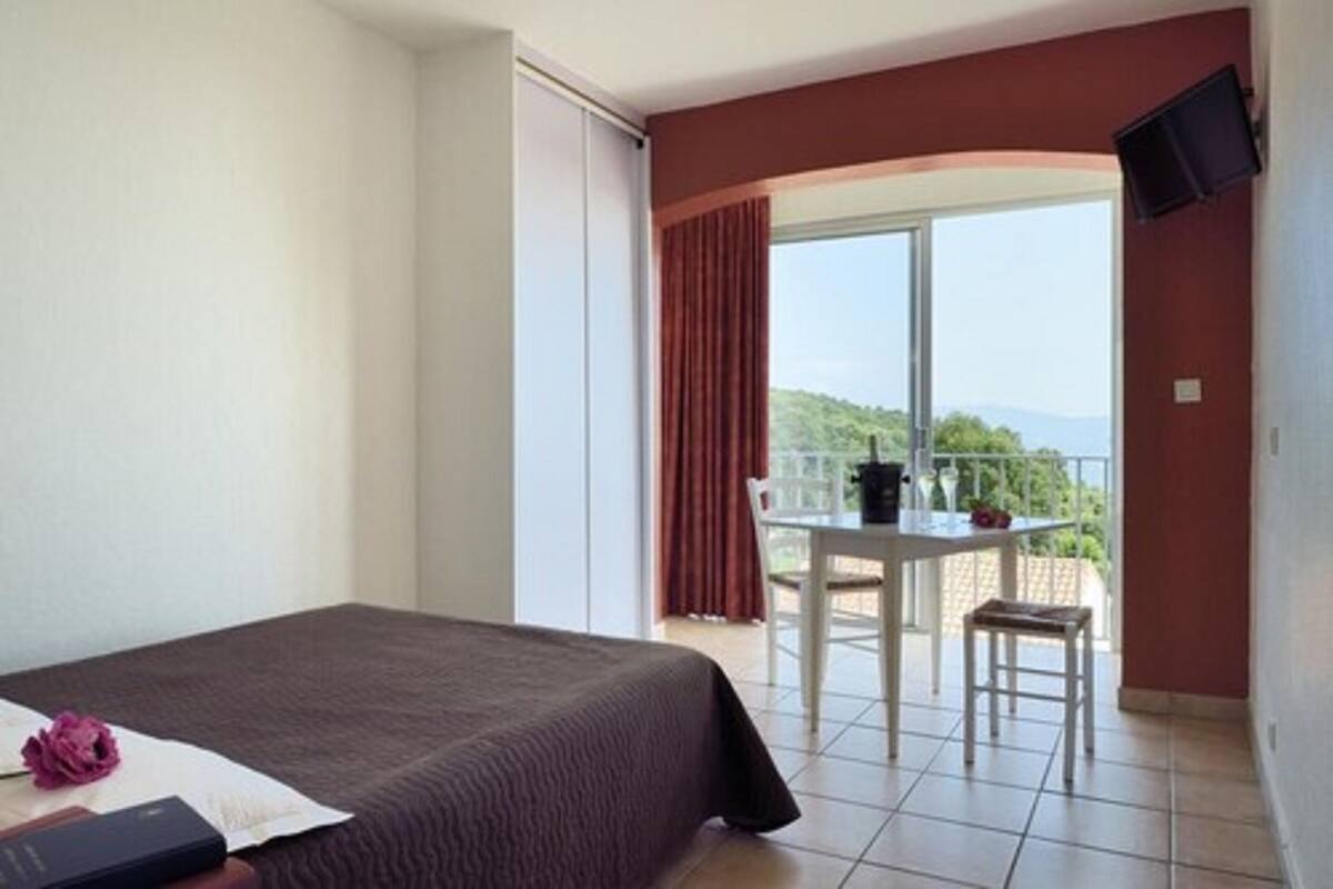 Une chambre d’hôtel avec un lit, une table et des chaises, et un balcon donnant sur un paysage verdoyant.