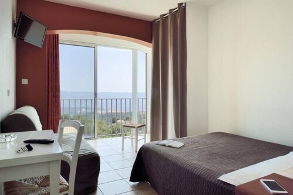 Une chambre d’hôtel comprenant un lit, un bureau avec une chaise, un téléviseur fixé au mur et un balcon avec des rideaux.