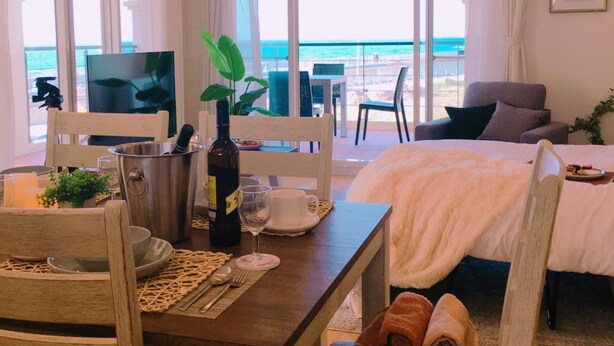 A hotel room with a bed, a dining table with chairs, a TV, and a view of the ocean. - Villa Del Oro Oceanside (Chatan)