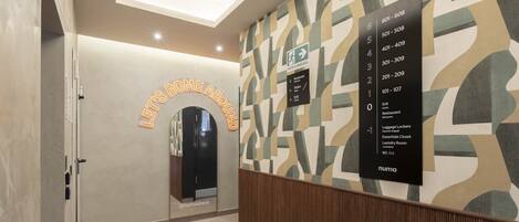 Un hall d'hôtel avec une pancarte indiquant « Bienvenue chez nous », un espace pour déposer les bagages et un mur affichant les numéros des chambres et les indications pour s'y rendre.
