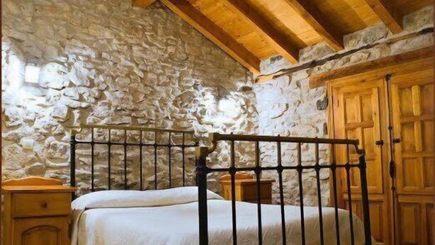 A bedroom with a stone wall, wooden ceiling beams, a metal bed frame, and a wooden wardrobe. - Casa Rural Las Trojes (Tamajon)
