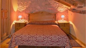 A bedroom with a wooden ceiling, stone wall, a bed with a patterned bedspread, two bedside tables with lamps, and a patterned rug on the floor. - Casa Rural Las Trojes (Tamajon)