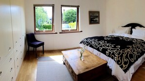 A bedroom with a bed, a chair, a wooden table, and two windows with a view of greenery. - The Eco-Flat (Sherborne)