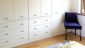 A room with a large white wardrobe, a dark blue chair, and a wooden coffee table with a vase of white flowers. - The Eco-Flat (Sherborne)