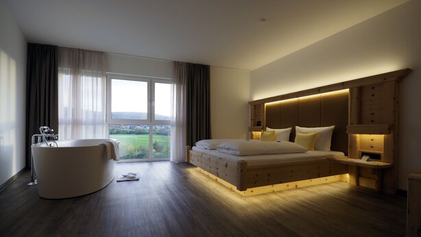 A modern hotel room with a large bed, a separate bathtub, and a view of a green landscape through the windows. - Hotel Reich an der Rems (Schorndorf)