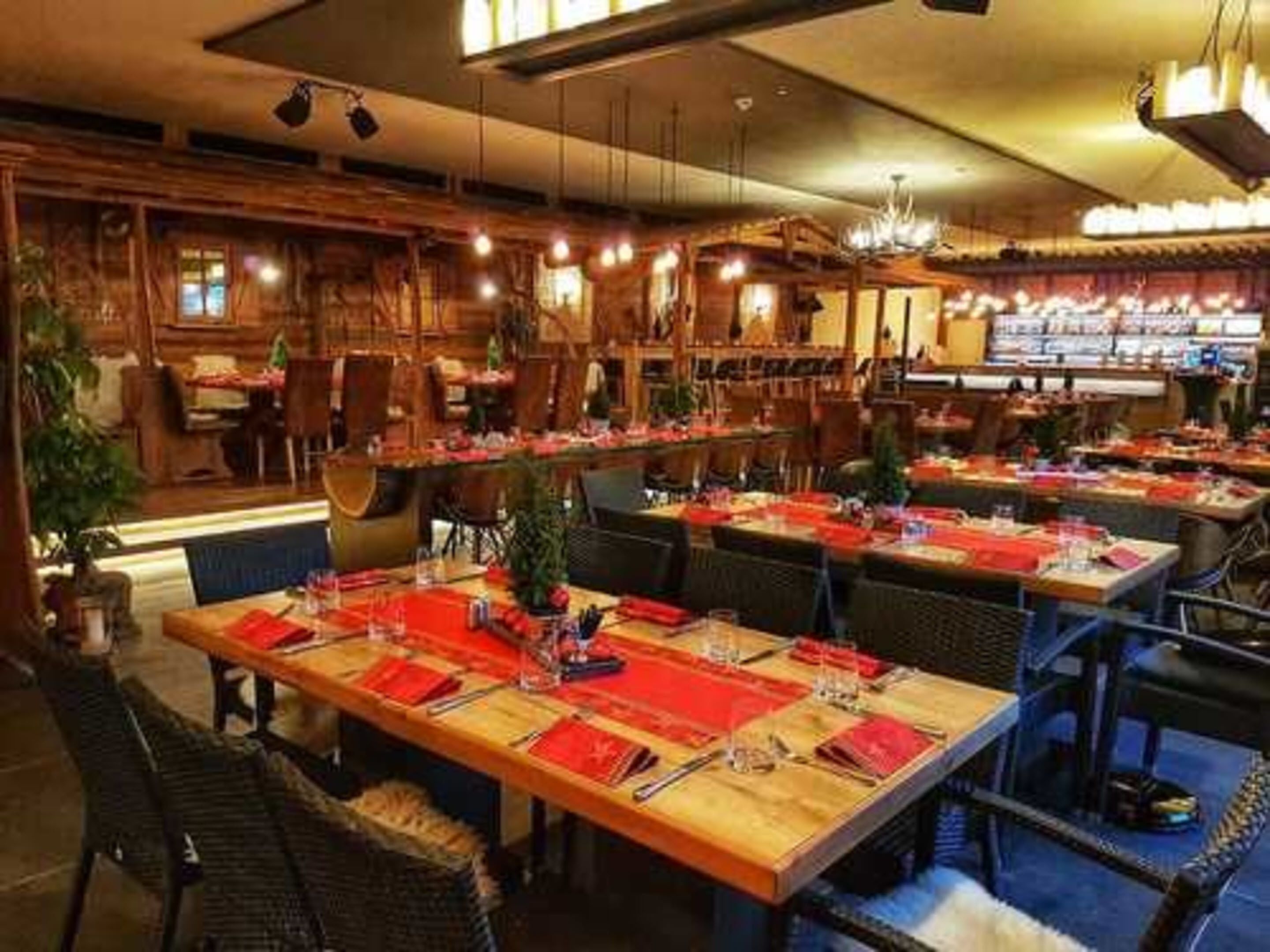 Ein Restaurant mit mehreren Esstischen, gedeckt mit roten Platzsets und Besteck, Holzstühlen und hängenden Lampen.