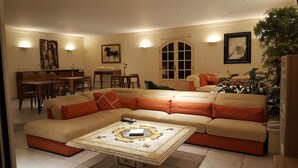 Ein geräumiges Wohnzimmer mit einer großen, beigefarbenen Eckcouch, einem passenden Hocker und einem dekorativen Couchtisch. Es gibt einen Essbereich mit Holzmöbeln und ein großes Fenster mit Blick.