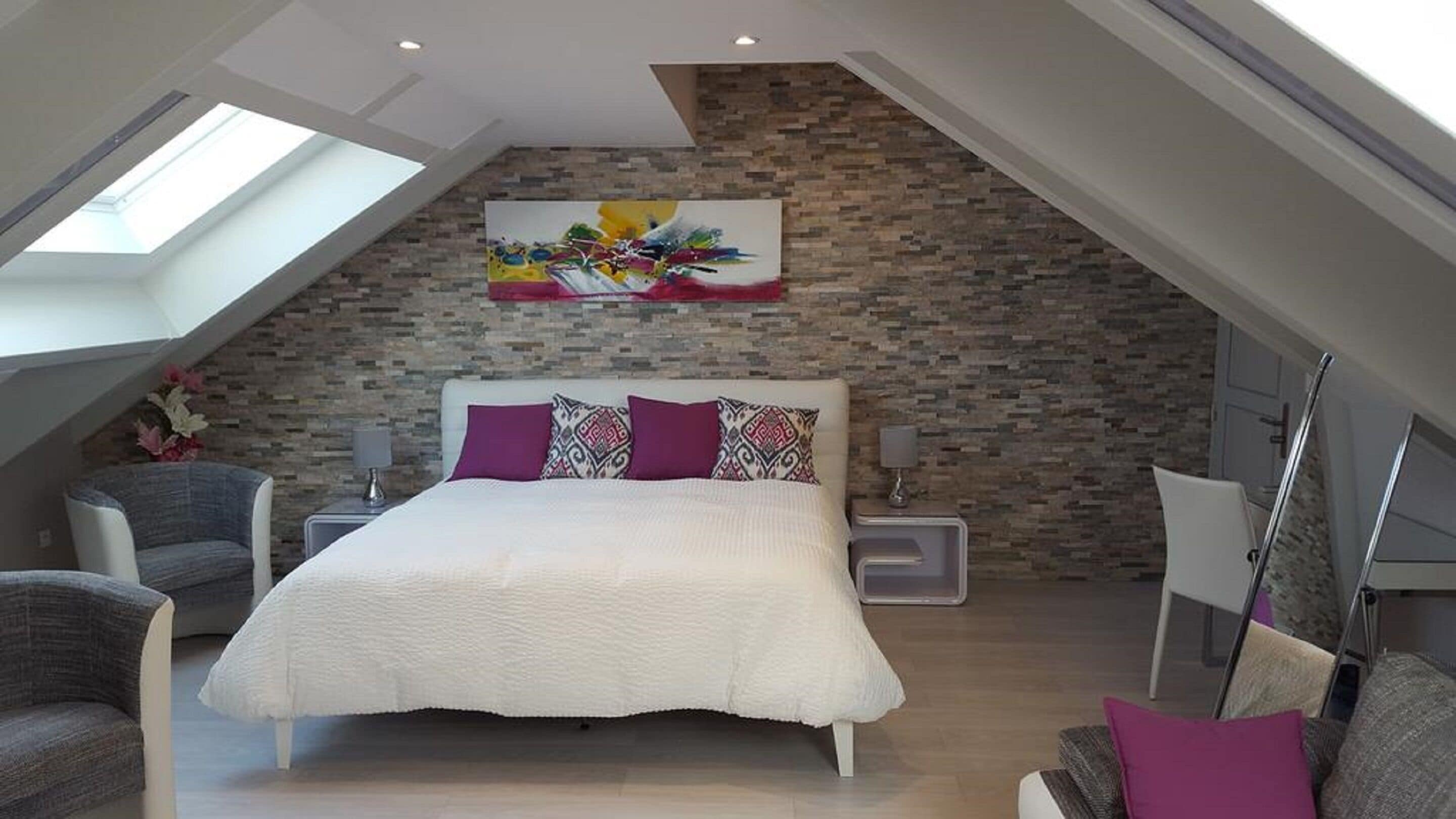 Un dormitorio moderno con techo inclinado, una cama grande con almohadones estampados, una pared de acento de piedra y un tragaluz.
