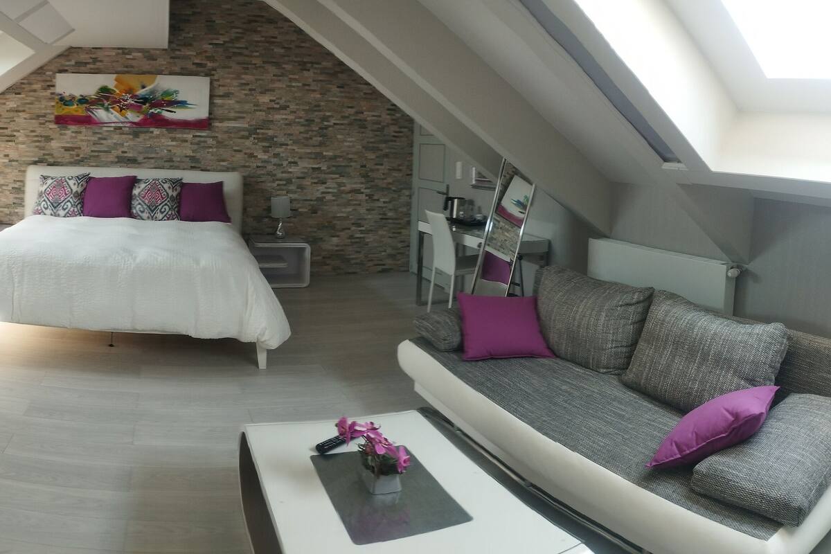 Une chambre de style loft moderne, avec un plafond en pente, un lit avec une literie blanche et des coussins violets, un canapé gris avec un coussin violet, une petite table avec un vase et un mur en briques décoré d’un tableau coloré.