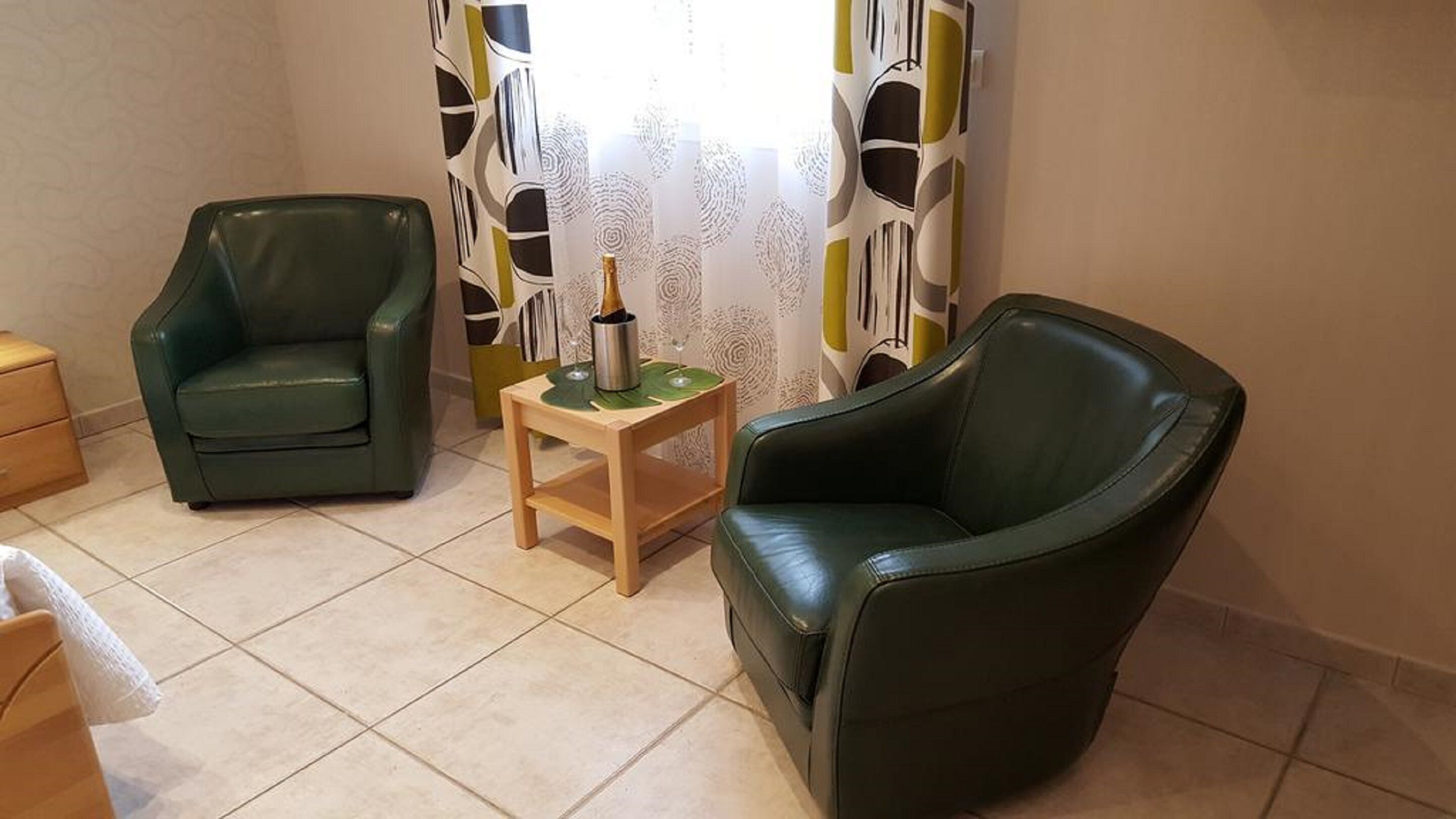 A room with two green leather chairs, a small wooden table with a bottle and glass, and patterned curtains.
