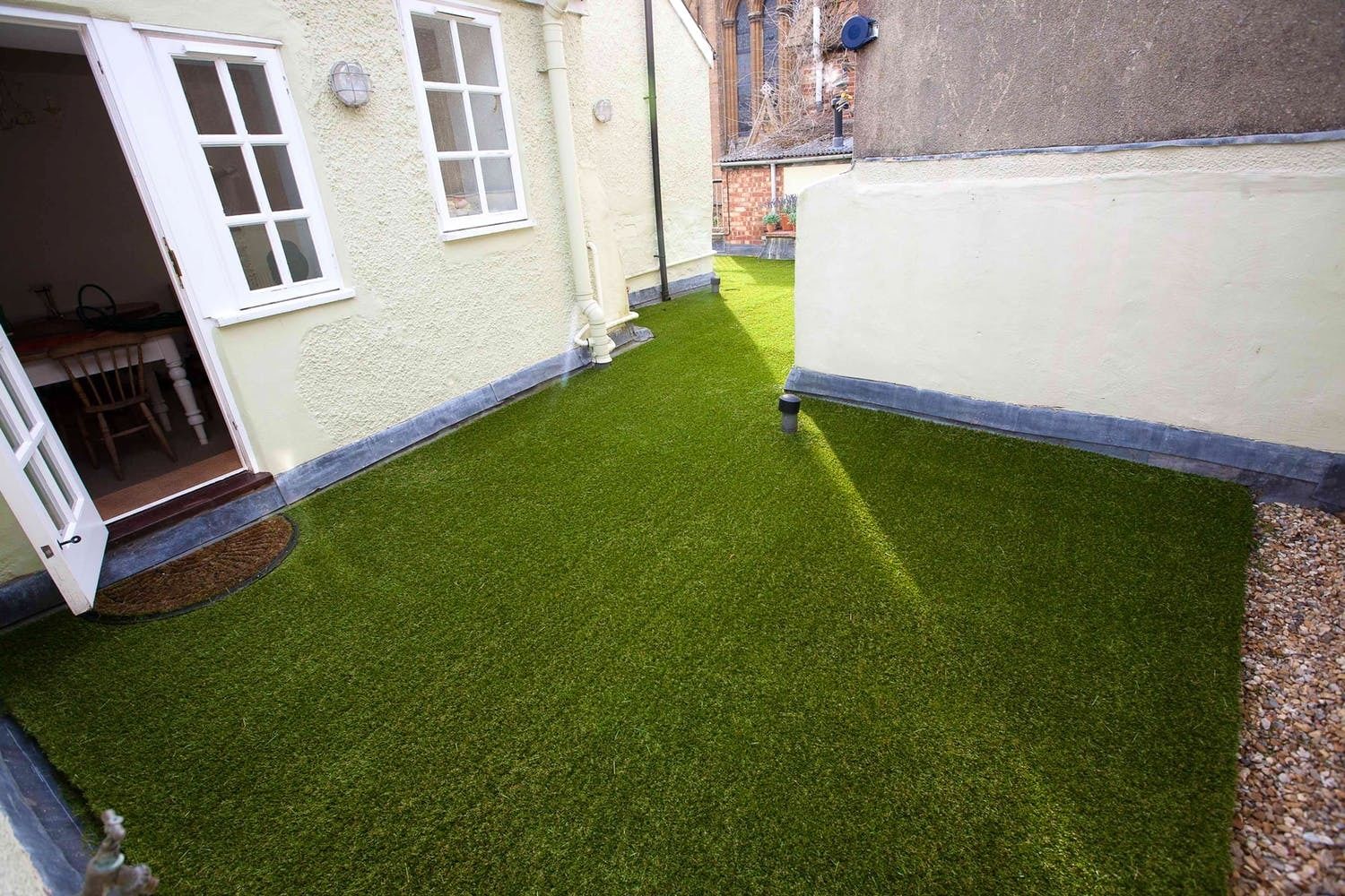 A narrow courtyard with artificial green grass, white walls, and a door leading to a room with a chair and a table.