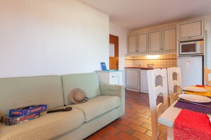 Une cuisine compacte avec des meubles blancs, un four à micro-ondes et un réfrigérateur. On y trouve une table à manger avec un chemin de table rouge et des assiettes blanches. Un canapé beige, agrémenté d'un chapeau et d'une télécommande, se trouve dans l'espace salon.