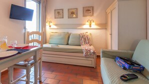 A cozy room with a bed, a TV mounted on the wall, a table with a glass pitcher and plates, and a couch with a game box on it. - Résidence Pierre & Vacances Baie de Saint-Tropez - Eden Parc (Saint-Tropez)